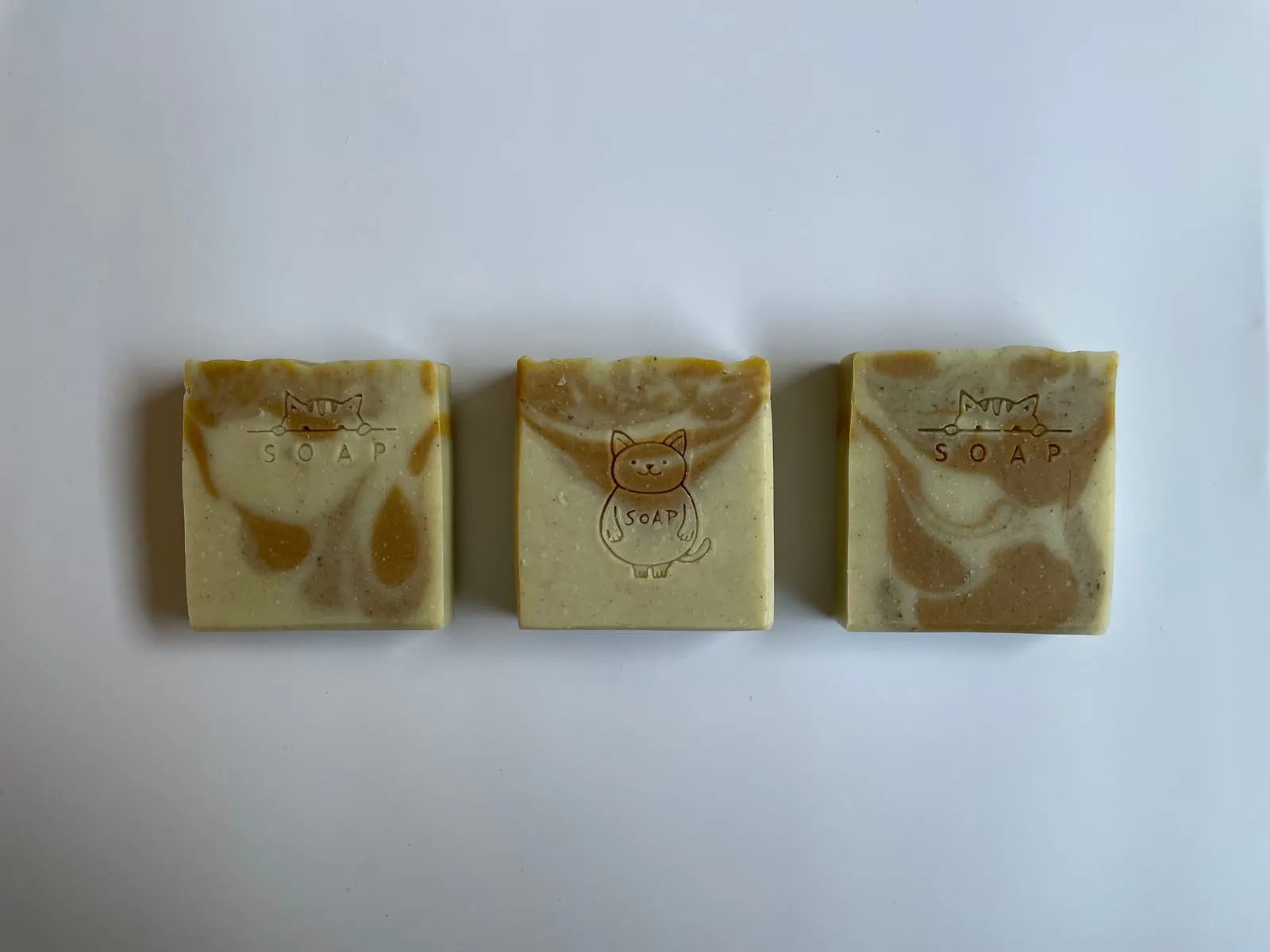 Three cold-process soap bars with cat stamp on the top, in a creamy yellow recipe with a touch of warm-orange swirl — a typical 5% superfat bar made from a coconut, olive, and shea butter blend.