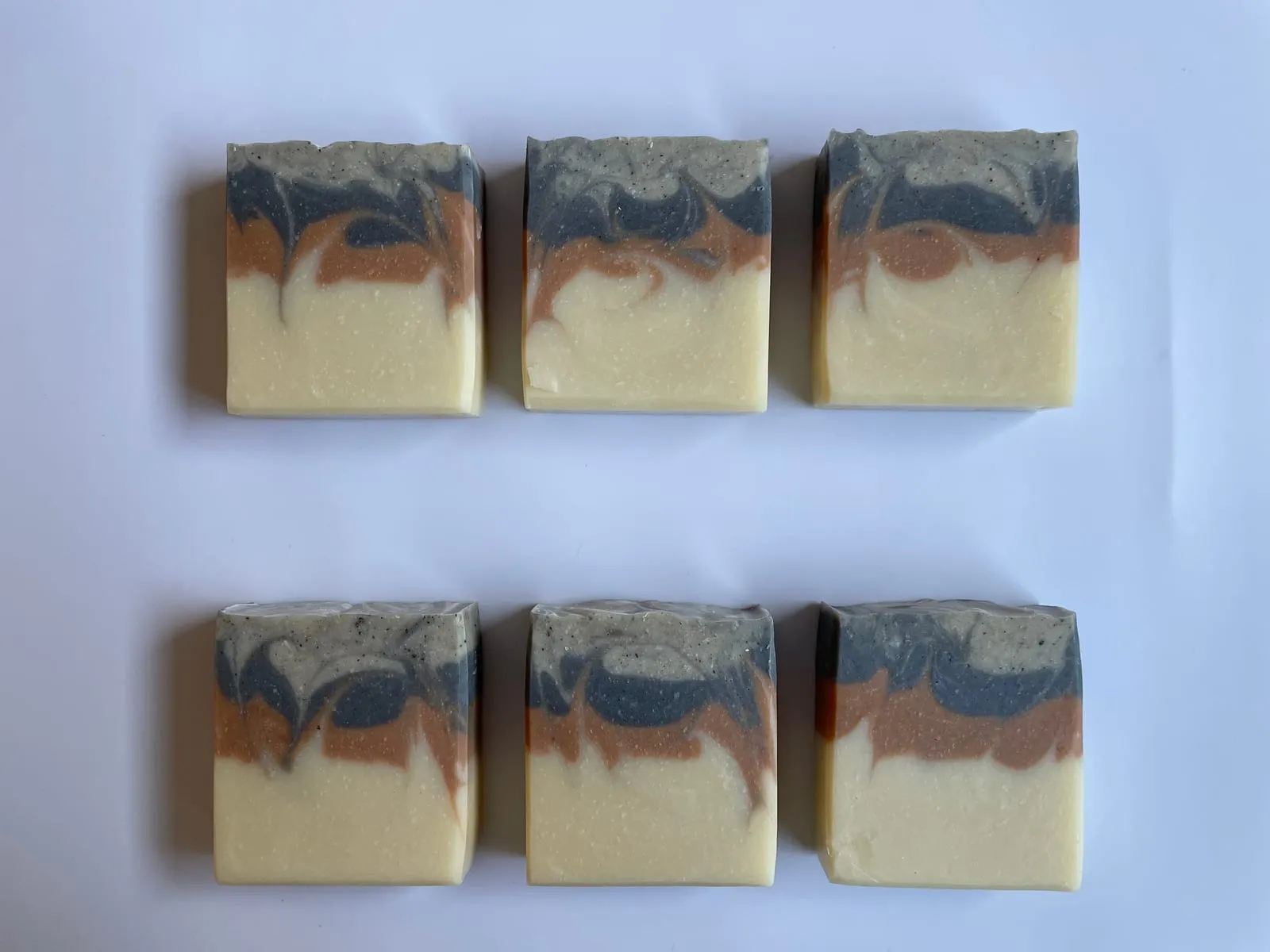Six cold-process soap bars laid in two rows of three, showing a clean horizontal three-band design — cream base, terracotta middle, grey-and-white swirled top. A 5% superfat recipe with reserved shea butter added at trace.